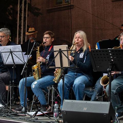 Auf der Tribüne sind Musiker der Bigband sitzend auf der Tribüne zu sehen. In der ersten Reihe, im Vordergrund, die Saxofonisten.