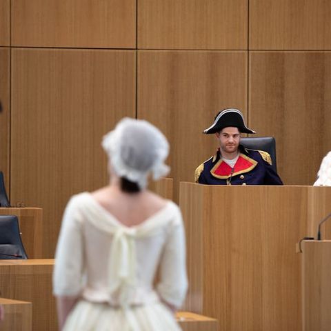 Auf dem Bild sieht man eine szenische Aufführung im Plenarsaal des Landtags Rheinland-Pfalz. Im Vordergrund stehen zwei Schauspieler in historischen Kostümen:  Der männliche Darsteller trägt einen grünen Gehrock mit goldenem Hemd und Weste.  Die weibliche Darstellerin ist in einem weißen Kleid mit Spitzenhaube gekleidet.  Im Hintergrund sitzen zwei weitere Darsteller am Rednerpult.