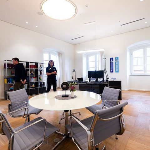 Auf dem Bild sieht man das Direktorenzimmer im Landtag Rheinland-Pfalz. Der Raum ist hell und freundlich gestaltet, mit großen Fenstern und eleganten Holzböden.  In der Mitte steht ein runder Besprechungstisch mit mehreren stilvollen Stühlen aus Metall und Stoff. Auf dem Tisch befindet sich eine kleine Blumenvase und ein dekorativer Untersetzer.  Im Hintergrund ist ein Schreibtisch mit zwei Monitoren zu sehen, daneben einige Dekorationsgegenstände und persönliche Elemente.  Links im Raum steht ein offenes Regal mit Büchern und bunten Objekten. An der Wand hängen zwei gerahmte Schilder/Banner.