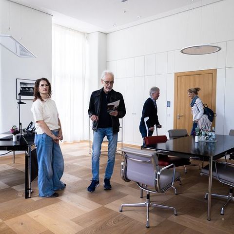 Auf dem Bild sieht man eine Gruppe von Personen im Präsidentenzimmer des Landtags Rheinland-Pfalz. Der Raum ist hell und großzügig gestaltet, mit großen Fenstern, die viel Tageslicht hereinlassen.  Auf der linken Seite steht ein eleganter Schreibtisch mit einem schwarzen Drehstuhl, einer Blumenvase und einem Telefon.  In der Mitte befindet sich ein großer Konferenztisch mit mehreren modernen Stühlen, auf dem Tisch stehen Wasserkaraffen und Gläser.  An der Wand sind große Schranktüren in einem minimalistischen Design zu sehen.