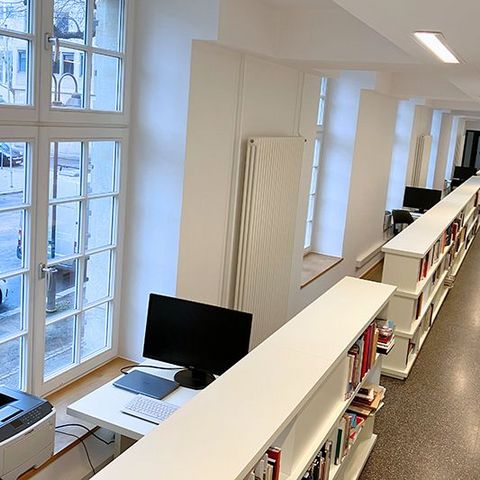 In einem langen Flur stehen mehrere aneinandergereihte Bücherregale. Neben ihnen befinden sich große Fenster und einzelne Tische mit Computermonitoren.