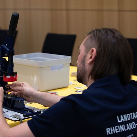 Auf dem Bild sieht man einen Mann, der eine Buttonmaschine bedient. Der Mann trägt ein dunkelblaues T-Shirt mit der Aufschrift 