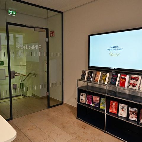 An einer Wand hängt ein Bildschirm mit der Aufschrift „Landtag Rheinland-Pfalz“ neben einer Glastür. Darunter steht ein Regal mit mehreren Büchern. Gegenüber befindet sich ein Tresen.