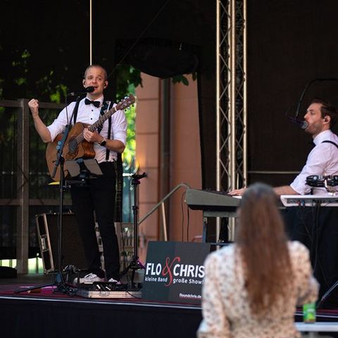 Auf dem Bild sieht man eine Live-Bühnenperformance des Duos Flo & Chris.  Bühne und Künstler Links auf der Bühne steht ein Musiker mit einer Gitarre, gekleidet in einem weißen Hemd mit schwarzer Fliege und Hosenträgern. Er hat ein Headset-Mikrofon und spricht oder singt ins Mikrofon.  Rechts sitzt ein weiterer Musiker an einem Keyboard, ebenfalls in einem weißen Hemd mit schwarzer Fliege und Hosenträgern, und singt in ein Mikrofon.