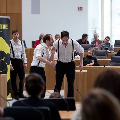Auf dem Bild sieht man eine Gruppe von Schauspielern, die eine Improvisationstheater-Aufführung im Plenarsaal des Landtags Rheinland-Pfalz präsentieren. Die Schauspieler tragen weiße Hemden mit schwarzen Hosenträgern und schwarzen Hosen. Zwei männliche Schauspieler stehen im Vordergrund und agieren miteinander in einer lebhaften Szene, wobei einer dem anderen etwas ins Ohr flüstert.  Im Hintergrund stehen weitere Ensemblemitglieder, die das Geschehen beobachten und auf ihren Einsatz warten.  Links neben der Bühne steht ein großes Plakat mit der Aufschrift 