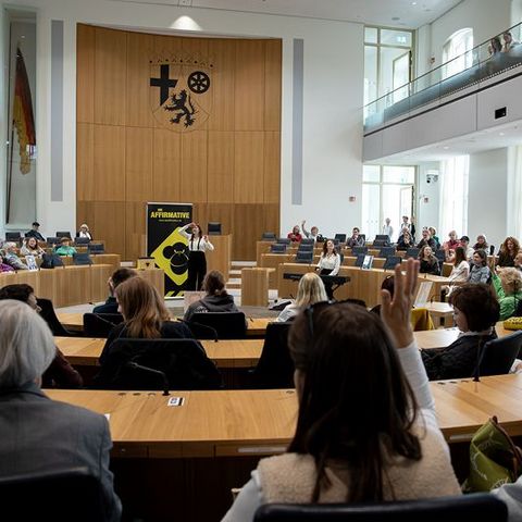 Zu sehen ist der Plenarsaal des Landtags Rheinland-Pfalz. Im Vordergrund stehen viele Menschen. Im Zentrum steht eine Person an einem Rednerpult. Hinter dieser Person ist ein auffälliges gelb-schwarzes Plakat mit der Aufschrift 