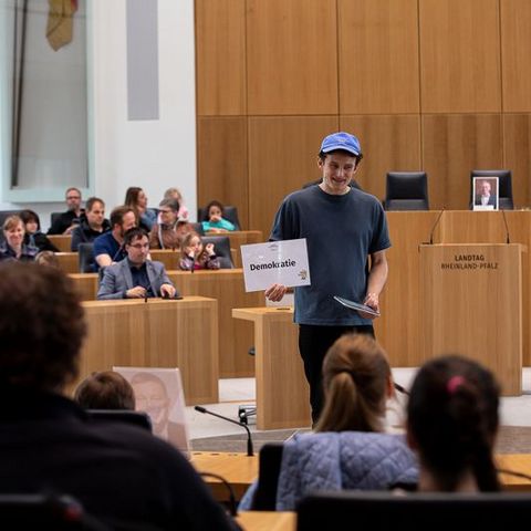 Auf dem Bild sieht man eine Veranstaltung im Plenarsaal des Landtags Rheinland-Pfalz. Im Vordergrund steht ein Mann mit einer blauen Kappe und einem dunkelblauen T-Shirt. Er hält ein Schild mit der Aufschrift 