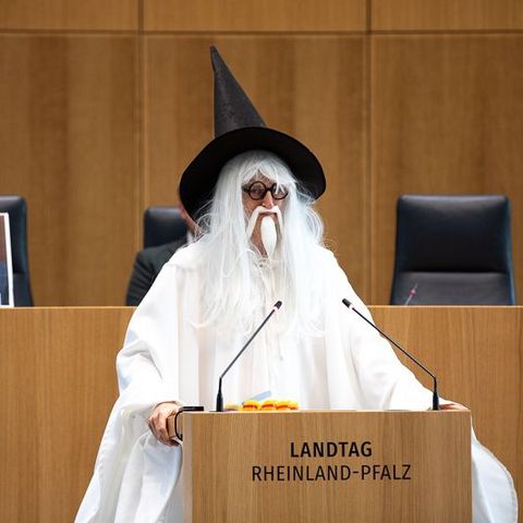 Auf dem Bild sieht man eine Person, die als Zauberer verkleidet ist. Die Person trägt ein langes weißes Gewand, eine weiße Perücke mit langem Haar und Bart sowie einen großen schwarzen Spitzhut. Sie steht hinter dem Rednerpult im Plenarsaal des Landtags Rheinland-Pfalz.