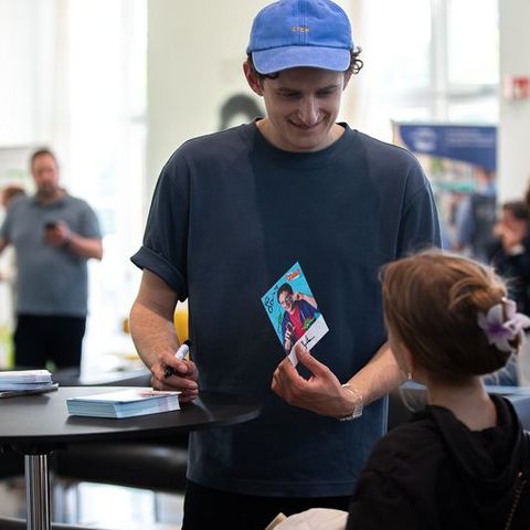 Auf dem Bild sieht man einen jungen Mann mit einer blauen Kappe und einem dunkelblauen T-Shirt. Er steht an einem Stehtisch und signiert Autogrammkarten für Kinder. Der Mann lächelt freundlich und hält eine Karte mit seinem Bildmotiv in der Hand.