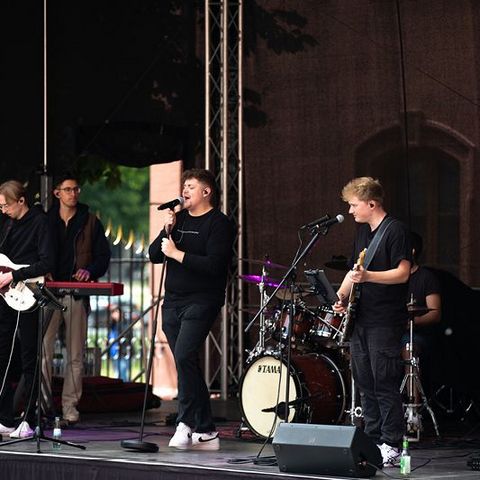 Auf dem Bild sieht man eine Live-Band, die auf einer Bühne während des Verfassungsfests 2025 im Freien auftritt. Die Band besteht aus mehreren jungen Musikern, die schwarze Kleidung tragen.  Im Vordergrund steht der Leadsänger mit einem Mikrofon, während ein Gitarrist rechts daneben ebenfalls in das Mikrofon singt.  Links auf der Bühne steht ein weiterer Gitarrist, daneben ein Keyboarder.  Im Hintergrund sitzt ein Schlagzeuger hinter einem Yamaha-Schlagzeug und begleitet den Auftritt rhythmisch.