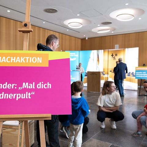 Auf dem Bild sieht man eine Mitmachaktion für Kinder im Rahmen einer Veranstaltung im Landtag Rheinland-Pfalz. Im Vordergrund steht ein buntes Schild auf einer Staffelei mit der Aufschrift: 