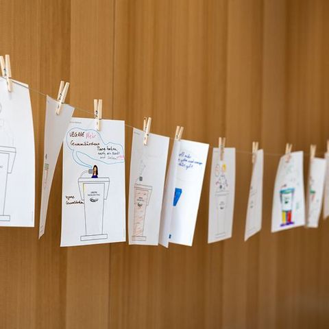 Auf dem Bild sieht man die Ergebnisse einer Mitmachaktion für Kinder im Rahmen einer Veranstaltung im Landtag Rheinland-Pfalz. Die Bilder sind auf einem dünnen Seil aufgehängt und mit kleinen Holzklammern befestigt.  Jedes Bild zeigt eine Zeichnung eines Rednerpults des Landtags Rheinland-Pfalz mit individuellen Ausgestaltungen.  Manche Zeichnungen sind farbig gestaltet, andere enthalten Sprechblasen oder kurze Botschaften.