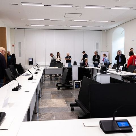 Auf dem Bild sieht man einen Sitzungssaal im Landtag Rheinland-Pfalz. Der Raum ist hell und modern gestaltet, mit großen Fenstern und weißen Wänden.  Die Sitzanordnung ist in U-Form angeordnet. Auf den Tischen stehen Mikrofone und Namensschilder, die die Positionen der Teilnehmer markieren.  Vor einigen Plätzen sind Porträtfotos aufgestellt, die die Position von bestimmten Abgeordneten darstellen.