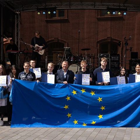 Auf dem Bild sieht man eine Gruppe von Personen, die vor einer Bühne stehen und eine große Europaflagge mit gelben Sternen auf blauem Hintergrund präsentieren.