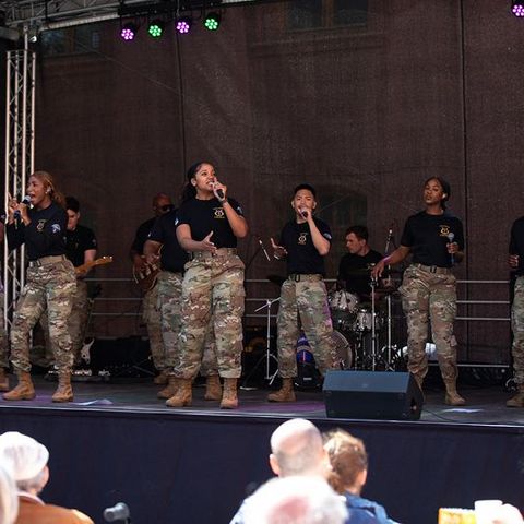 Auf dem Bild sieht man eine musikalische Aufführung auf einer Bühne im Freien. Die Gruppe besteht aus mehreren Sängerinnen und Sängern sowie Musikern, die Militäruniformen tragen. Es handelt sich um die USAREUR-AF Band and Chorus. Die Künstler:innen tragen schwarze T-Shirts mit einem Emblem und militärische Tarnhosen sowie Stiefel.  Die Frontsängerinnen und -sänger singen in Mikrofone, während die Band im Hintergrund spielt. Zu sehen sind unter anderem ein Schlagzeuger, ein Gitarrist und ein Keyboarder.