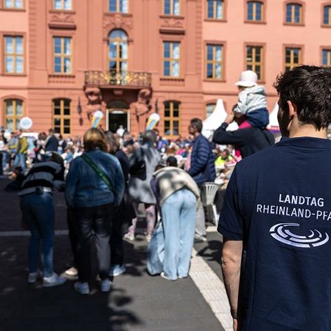 Im Vordergrund steht ein Mann mit einem dunkelblauen T-Shirt, auf dessen Rückseite die Aufschrift 