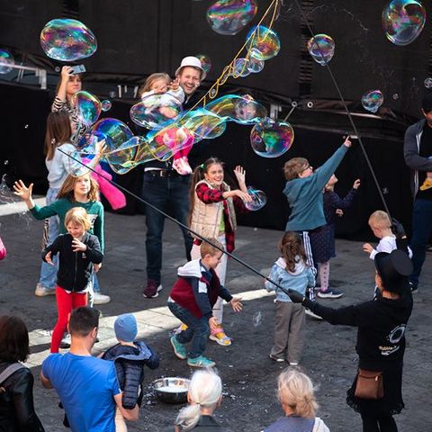 Auf dem Bild sieht man eine fröhliche Szene während des Verfassungsfests, bei der Kinder und Erwachsene mit großen Seifenblasen spielen. Im Mittelpunkt stehen Kinder, die begeistert nach großen Seifenblasen greifen und versuchen, diese zu fangen oder platzen zu lassen.  Eine Person, die zum Event-Team zu gehören scheint, produziert die Seifenblasen mit langen Stäben und Seifenlauge.  Erwachsene begleiten die Kinder und beobachten das Geschehen, einige halten Kleinkinder auf dem Arm.