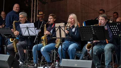 Auf der Tribüne sind Musiker der Bigband sitzend auf der Tribüne zu sehen. In der ersten Reihe, im Vordergrund, die Saxofonisten.