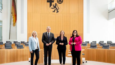 Zu sehen sind vier Personen nebeneinanderstehend im Rund des Plenarsaals. Im Hintergrund sieht man das Wappen von RLP, das Rednerpult und die Hambacher Fahne.