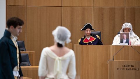 Auf dem Bild sieht man eine szenische Aufführung im Plenarsaal des Landtags Rheinland-Pfalz. Im Vordergrund stehen zwei Schauspieler in historischen Kostümen:  Der männliche Darsteller trägt einen grünen Gehrock mit goldenem Hemd und Weste.  Die weibliche Darstellerin ist in einem weißen Kleid mit Spitzenhaube gekleidet.  Im Hintergrund sitzen zwei weitere Darsteller am Rednerpult.