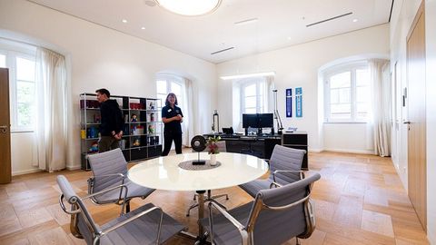 Auf dem Bild sieht man das Direktorenzimmer im Landtag Rheinland-Pfalz. Der Raum ist hell und freundlich gestaltet, mit großen Fenstern und eleganten Holzböden.  In der Mitte steht ein runder Besprechungstisch mit mehreren stilvollen Stühlen aus Metall und Stoff. Auf dem Tisch befindet sich eine kleine Blumenvase und ein dekorativer Untersetzer.  Im Hintergrund ist ein Schreibtisch mit zwei Monitoren zu sehen, daneben einige Dekorationsgegenstände und persönliche Elemente.  Links im Raum steht ein offenes Regal mit Büchern und bunten Objekten. An der Wand hängen zwei gerahmte Schilder/Banner.
