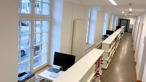 In einem langen Flur stehen mehrere aneinandergereihte Bücherregale. Neben ihnen befinden sich große Fenster und einzelne Tische mit Computermonitoren.