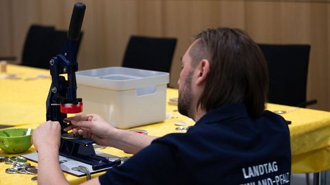 Auf dem Bild sieht man einen Mann, der eine Buttonmaschine bedient. Der Mann trägt ein dunkelblaues T-Shirt mit der Aufschrift 