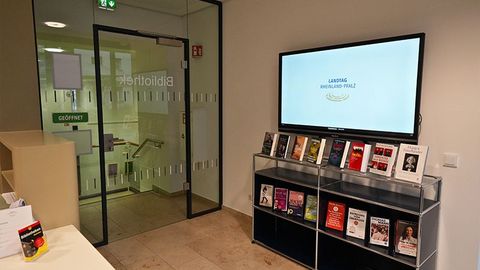 An einer Wand hängt ein Bildschirm mit der Aufschrift „Landtag Rheinland-Pfalz“ neben einer Glastür. Darunter steht ein Regal mit mehreren Büchern. Gegenüber befindet sich ein Tresen.