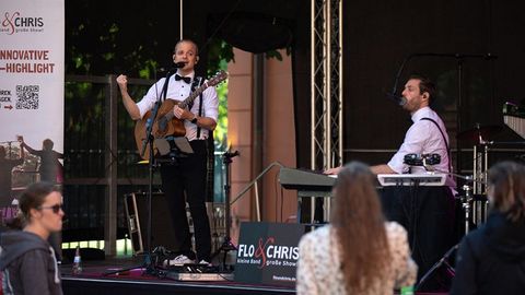 Auf dem Bild sieht man eine Live-Bühnenperformance des Duos Flo & Chris.  Bühne und Künstler Links auf der Bühne steht ein Musiker mit einer Gitarre, gekleidet in einem weißen Hemd mit schwarzer Fliege und Hosenträgern. Er hat ein Headset-Mikrofon und spricht oder singt ins Mikrofon.  Rechts sitzt ein weiterer Musiker an einem Keyboard, ebenfalls in einem weißen Hemd mit schwarzer Fliege und Hosenträgern, und singt in ein Mikrofon.