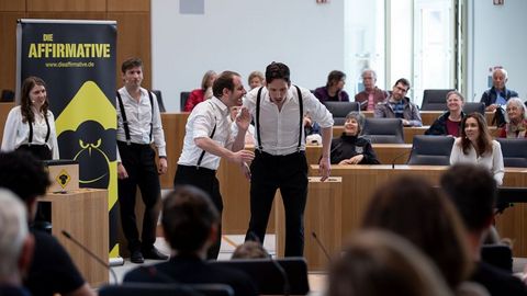 Auf dem Bild sieht man eine Gruppe von Schauspielern, die eine Improvisationstheater-Aufführung im Plenarsaal des Landtags Rheinland-Pfalz präsentieren. Die Schauspieler tragen weiße Hemden mit schwarzen Hosenträgern und schwarzen Hosen. Zwei männliche Schauspieler stehen im Vordergrund und agieren miteinander in einer lebhaften Szene, wobei einer dem anderen etwas ins Ohr flüstert.  Im Hintergrund stehen weitere Ensemblemitglieder, die das Geschehen beobachten und auf ihren Einsatz warten.  Links neben der Bühne steht ein großes Plakat mit der Aufschrift 