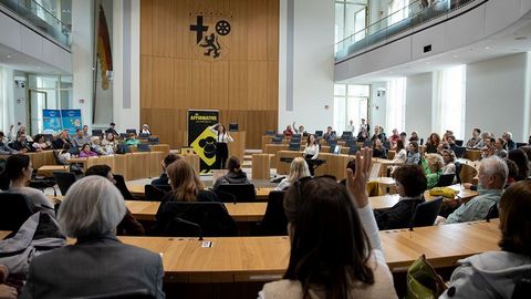 Zu sehen ist der Plenarsaal des Landtags Rheinland-Pfalz. Im Vordergrund stehen viele Menschen. Im Zentrum steht eine Person an einem Rednerpult. Hinter dieser Person ist ein auffälliges gelb-schwarzes Plakat mit der Aufschrift 