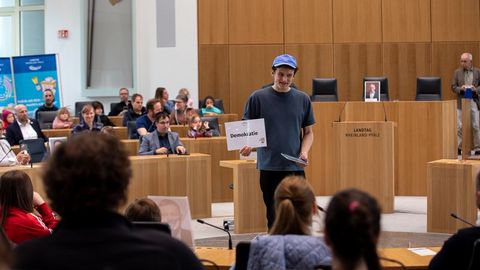 Auf dem Bild sieht man eine Veranstaltung im Plenarsaal des Landtags Rheinland-Pfalz. Im Vordergrund steht ein Mann mit einer blauen Kappe und einem dunkelblauen T-Shirt. Er hält ein Schild mit der Aufschrift 