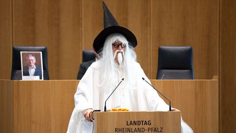 Auf dem Bild sieht man eine Person, die als Zauberer verkleidet ist. Die Person trägt ein langes weißes Gewand, eine weiße Perücke mit langem Haar und Bart sowie einen großen schwarzen Spitzhut. Sie steht hinter dem Rednerpult im Plenarsaal des Landtags Rheinland-Pfalz.