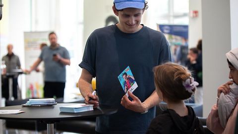 Auf dem Bild sieht man einen jungen Mann mit einer blauen Kappe und einem dunkelblauen T-Shirt. Er steht an einem Stehtisch und signiert Autogrammkarten für Kinder. Der Mann lächelt freundlich und hält eine Karte mit seinem Bildmotiv in der Hand.