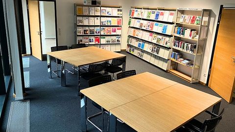 In der Mitte eines Raums stehen zwei rechteckige Holztische mit mehreren Stühlen. Im Hintergrund befinden sich Wandregale mit Zeitschriften und Büchern.