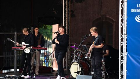 Auf dem Bild sieht man eine Live-Band, die auf einer Bühne während des Verfassungsfests 2025 im Freien auftritt. Die Band besteht aus mehreren jungen Musikern, die schwarze Kleidung tragen.  Im Vordergrund steht der Leadsänger mit einem Mikrofon, während ein Gitarrist rechts daneben ebenfalls in das Mikrofon singt.  Links auf der Bühne steht ein weiterer Gitarrist, daneben ein Keyboarder.  Im Hintergrund sitzt ein Schlagzeuger hinter einem Yamaha-Schlagzeug und begleitet den Auftritt rhythmisch.