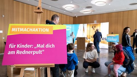 Auf dem Bild sieht man eine Mitmachaktion für Kinder im Rahmen einer Veranstaltung im Landtag Rheinland-Pfalz. Im Vordergrund steht ein buntes Schild auf einer Staffelei mit der Aufschrift: 