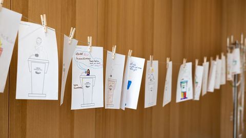 Auf dem Bild sieht man die Ergebnisse einer Mitmachaktion für Kinder im Rahmen einer Veranstaltung im Landtag Rheinland-Pfalz. Die Bilder sind auf einem dünnen Seil aufgehängt und mit kleinen Holzklammern befestigt.  Jedes Bild zeigt eine Zeichnung eines Rednerpults des Landtags Rheinland-Pfalz mit individuellen Ausgestaltungen.  Manche Zeichnungen sind farbig gestaltet, andere enthalten Sprechblasen oder kurze Botschaften.