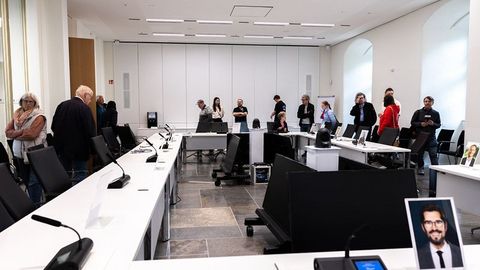 Auf dem Bild sieht man einen Sitzungssaal im Landtag Rheinland-Pfalz. Der Raum ist hell und modern gestaltet, mit großen Fenstern und weißen Wänden.  Die Sitzanordnung ist in U-Form angeordnet. Auf den Tischen stehen Mikrofone und Namensschilder, die die Positionen der Teilnehmer markieren.  Vor einigen Plätzen sind Porträtfotos aufgestellt, die die Position von bestimmten Abgeordneten darstellen.