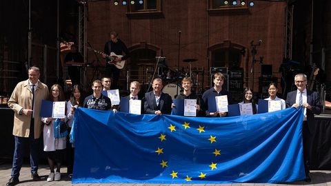 Auf dem Bild sieht man eine Gruppe von Personen, die vor einer Bühne stehen und eine große Europaflagge mit gelben Sternen auf blauem Hintergrund präsentieren.