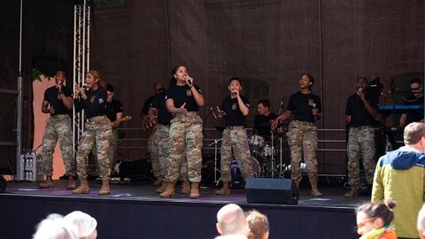 Auf dem Bild sieht man eine musikalische Aufführung auf einer Bühne im Freien. Die Gruppe besteht aus mehreren Sängerinnen und Sängern sowie Musikern, die Militäruniformen tragen. Es handelt sich um die USAREUR-AF Band and Chorus. Die Künstler:innen tragen schwarze T-Shirts mit einem Emblem und militärische Tarnhosen sowie Stiefel.  Die Frontsängerinnen und -sänger singen in Mikrofone, während die Band im Hintergrund spielt. Zu sehen sind unter anderem ein Schlagzeuger, ein Gitarrist und ein Keyboarder.