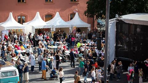 Auf dem Bild sieht man eine große Menschenmenge auf einem Platz vor dem Gebäude des Landtags Rheinland-Pfalz. Im Vordergrund stehen zahlreiche Menschen, die sich um Stehtische gruppieren oder durch die Menge schlendern. Einige sitzen an Tischen unter weißen Pavillons, die als Infostände oder Verpflegungsstände dienen.  Auf der rechten Seite sieht man eine Bühne. Die weißen Pagodenzelte sind festlich geschmückt, und es hängen bunte Girlanden sowie Banner.  Im linken Vordergrund parkt ein türkisfarbener VW-Bus.