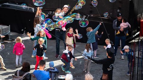 Auf dem Bild sieht man eine fröhliche Szene während des Verfassungsfests, bei der Kinder und Erwachsene mit großen Seifenblasen spielen. Im Mittelpunkt stehen Kinder, die begeistert nach großen Seifenblasen greifen und versuchen, diese zu fangen oder platzen zu lassen.  Eine Person, die zum Event-Team zu gehören scheint, produziert die Seifenblasen mit langen Stäben und Seifenlauge.  Erwachsene begleiten die Kinder und beobachten das Geschehen, einige halten Kleinkinder auf dem Arm.