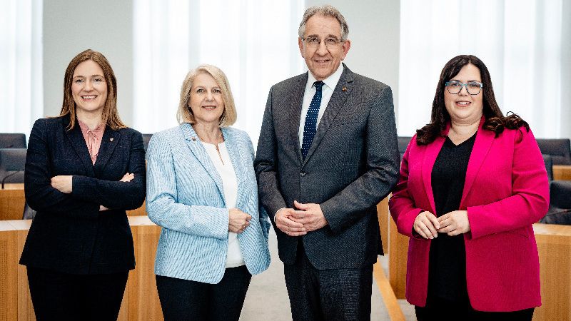 Zu sehen sind vier Personen nebeneinanderstehend im Rund des Plenarsaals.