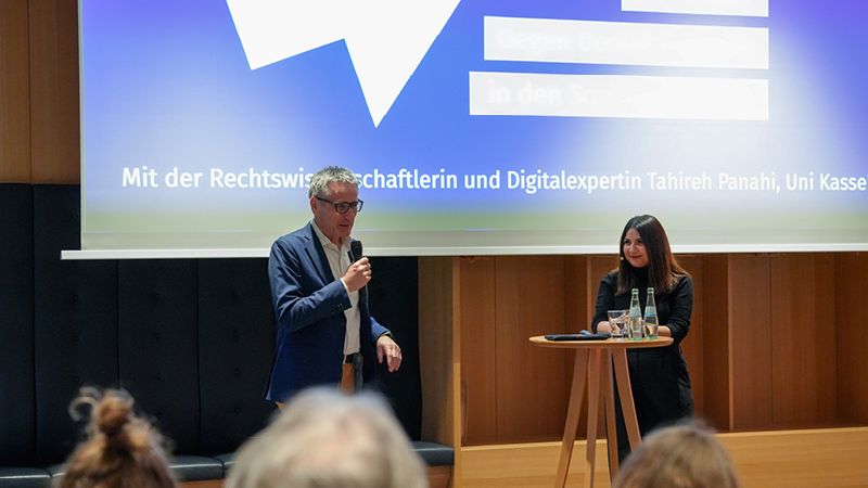 Zu sehen ist ein Mann und eine Frau. Sie stehen im Esszimmer des Landtags vor einer Leinwand. Auf der blauen Leinwand sieht man das Logo der Veranstaltungsreihe "Werte. Haltung. Demokratie.".