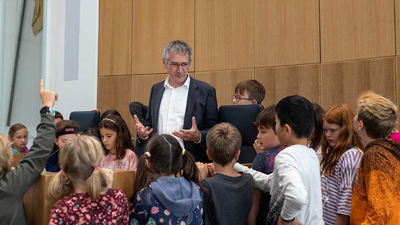 Auf dem Bild sieht man Landtagspräsident Hendrik Hering umringt von Schülerinnen und Schülern einer Grundschulklasse im Rahmen des Grundschulprogramms im Landtag, hier im Plenarsaaal.