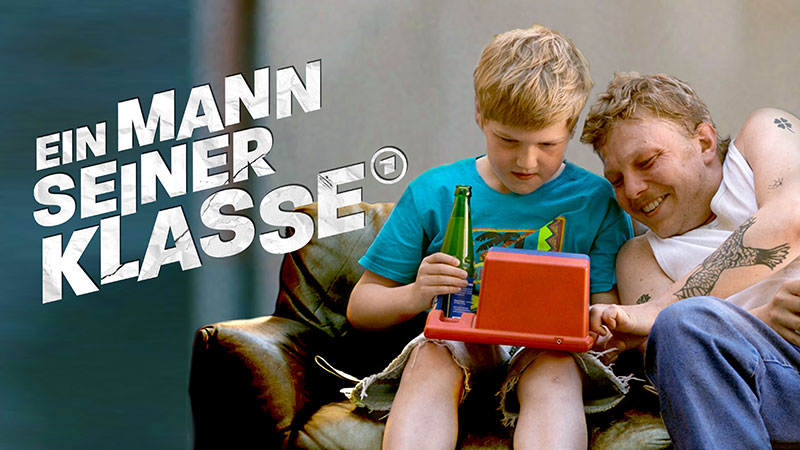 Vater und Sohn sitzen gemeinsam freudestrahlend auf einer Couch. Der hat ein Spielzeug auf dem Schoß. Das Bild trägt den Titel des Films: 