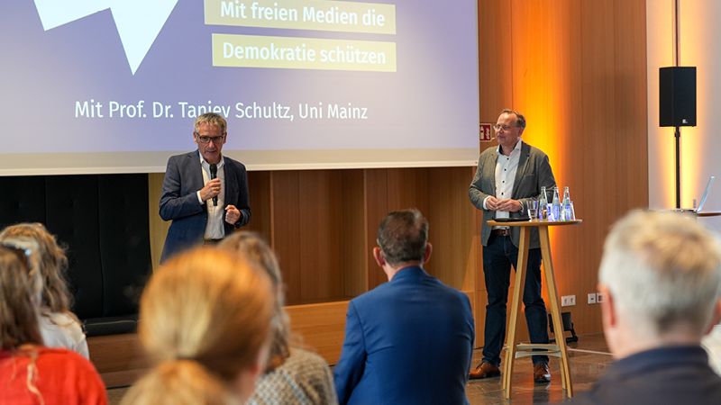 Zu sehen sind zwei M&auml;nner. Sie stehen im Restaurant des Landtags vor einer gro&szlig;en Leinwand. Auf der Leinwand steht: "#4 Tu was! Mit freien Medien die Demokratie sch&uuml;tzen, mit Prof. Dr. Tanjev Schultz". Im Vordergrund sitzt das Publikum. Der Mann auf der rechten Seite steht hinter einem Stehtisch. Auf diesem stehen Wasserflaschen. Der Mann auf der linken Seite h&auml;lt ein Mikrofon in der Hand und spricht zum Publikum.