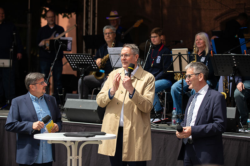 Der Ministerpräsident spricht in ein Mikrofon. Der Landtagspräsident und der Leiter der Blue Light Big Band des Polizeipräsidiums Ludwigshafen stehen neben ihm.
