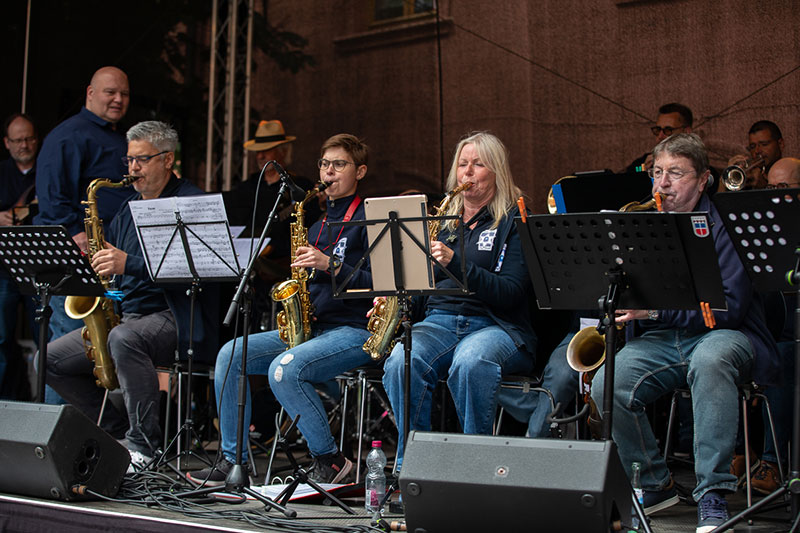 Auf der Tribüne sind Musiker der Bigband sitzend auf der Tribüne zu sehen. In der ersten Reihe, im Vordergrund, die Saxofonisten.